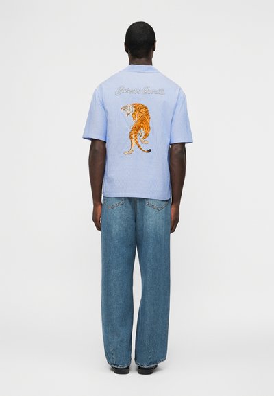 Άνδρας που φοράει ανοιχτό μπλε κοντομάνικο πουκάμισο με κεντημένη τίγρη και την επιγραφή «Roberto Cavalli» στην πλάτη, συνδυασμένο με φαρδιά μπλε τζιν και μαύρα παπούτσια.