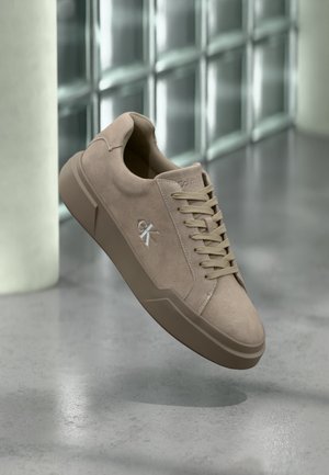 Baskets en daim beige avec lacets et logo "ck", présentées en suspension au-dessus d'un sol en béton poli avec un mur en blocs de verre en arrière-plan.