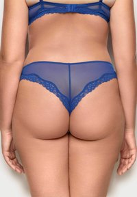 Modré krajkové a síťované thong spodní prádlo s vlnitými okraji, s průhlednou texturou a nízkým pasem pro minimální zakrytí.