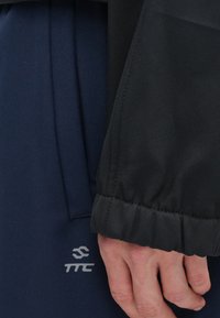 Μαύρο μανίκι από τζάκετ με ελαστική μανσέτα, επικαλυπτόμενο μπλε υφαντό ύφασμα. Γκρι λογότυπο τυπωμένο σε παντελόνι. Κοντινή λήψη, χωρίς στοιχεία φόντου.