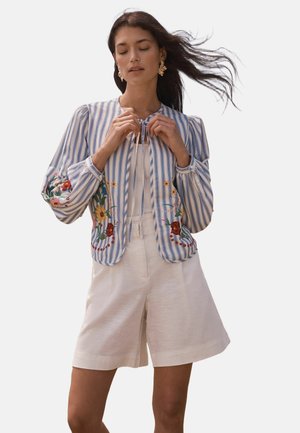 Woman with long dark hair wearing gold earrings, a blue-and-white striped floral embroidered jacket, and white high-waisted shorts.