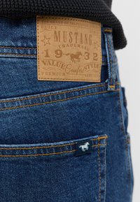 Jeans in denim con una toppa in pelle beige con testo impressa e logo di un cavallo. Dettagli di cucitura in oro, tasca posteriore con piccolo etichetta logo.