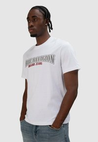 Witte katoenen T-shirt met een afbeelding van "True Religion Brand Jeans" in grijs en rood. Korte mouwen en een losse pasvorm, gedragen met lichtblauwe spijkerbroeken.