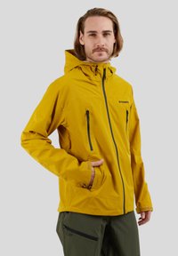 Gelbe wasserdichte Jacke mit Kapuze, ausgestattet mit Reißverschlusstaschen und glatter Textur. Das Model steht mit den Händen in den Taschen und trägt grüne Hosen.