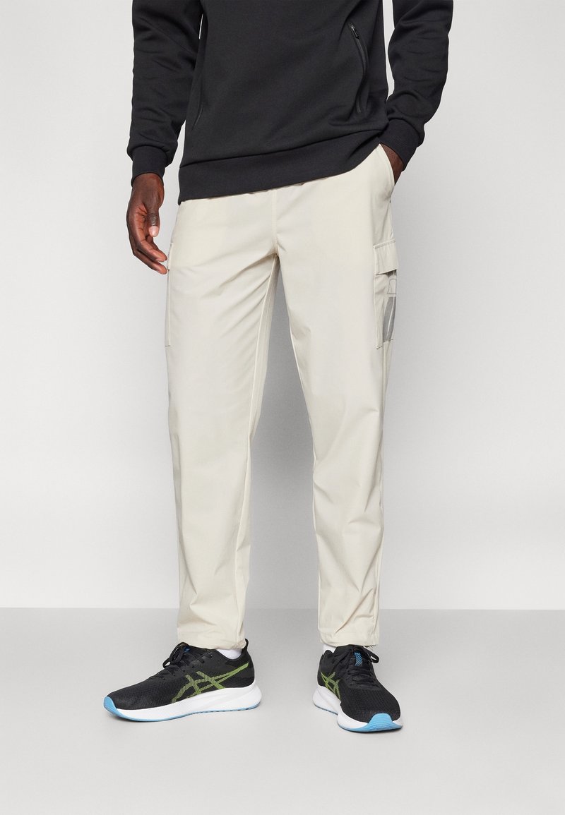 Pantalon cargo beige avec poches latérales, associé à des chaussures de course noires avec des accents verts et une semelle bleu clair. Sweat-shirt noir en haut.