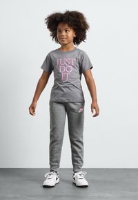 T-shirt cinza com "JUST DO IT." em rosa, combinado com calças de moletom cinzas com um logo da Nike em rosa. Ténis brancos completam o conjunto.