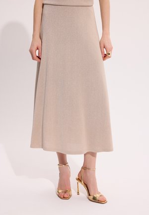 Kobieta w beżowej spódnicy midi, nosząca złote sandały na wysokim obcasie i złoty pierścionek na prawej ręce, stojąca na tle o jednolitym kolorze.