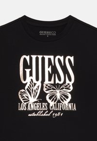 Black organic cotton t-shirt featuring large white text "GUESS" above two butterfly graphics and "LOS ANGELES CALIFORNIA established 1981."