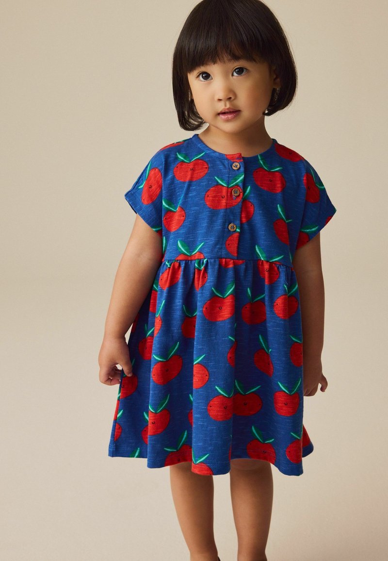 Young child with short black hair wearing a blue dress decorated with red smiling apple patterns, standing against a plain beige background.