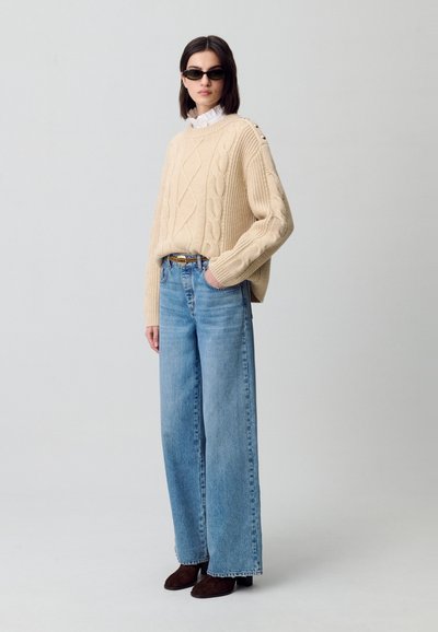 Maglione beige a trecce con bottoni sulle spalle, abbinato a pantaloni a gamba larga blu chiaro e stivali neri alla caviglia. Occhiali da sole neri completano l'outfit.