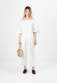 Blusa bianca con spalle scoperte e maniche a palloncino, decorata con un bordo floreale, abbinata a pantaloni bianchi a gamba larga e una piccola borsa strutturata. Scarpe nere.