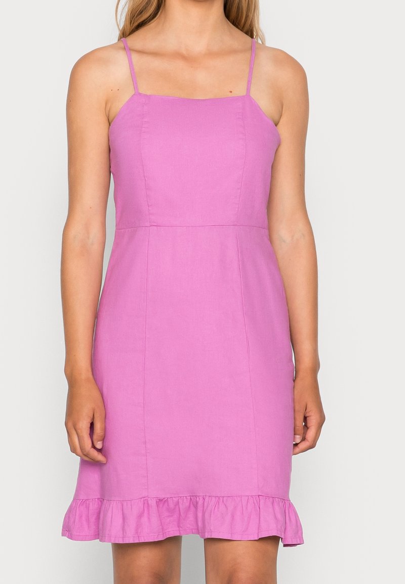 Femme portant une robe rose vif sans manches, longueur genou, avec de fines bretelles et un ourlet à volants, sur un fond clair uni.