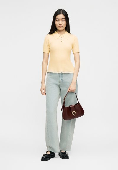 Νεαρή γυναίκα στέκεται φορώντας ένα απαλό κίτρινο πλεκτό τοπ με κοντά μανίκια, ανοιχτό μπλε τζιν παντελόνι με φαρδιά γραμμή, μαύρα μοκασίνια και κρατώντας μια σκούρα κόκκινη τσάντα.