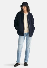 Navy bomber jacket with zip closure, layered over a cream sweatshirt. Light blue patchwork jeans and black shoes complete the outfit.