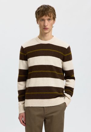 Jeune homme portant un pull rayé crème et marron avec de fines lignes jaunes et un pantalon kaki, debout devant un fond clair uni.