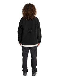 Person med krøllet hår stående iført en sort fleecejakke med teksten "ATW" på ryggen, sorte bukser, grå sko og en hvid skjorte under.