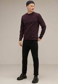 Felpa bordeaux con la scritta "GMTS" in rilievo sul davanti, abbinata a jeans neri e stivali neri con lacci, completata da un berretto scuro.