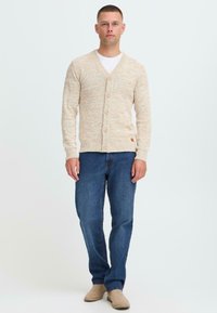 Mann som står, iført beige knappet cardigan, hvit skjorte, blå jeans og beige gamasjer støvler mot en ensfarget hvit bakgrunn.