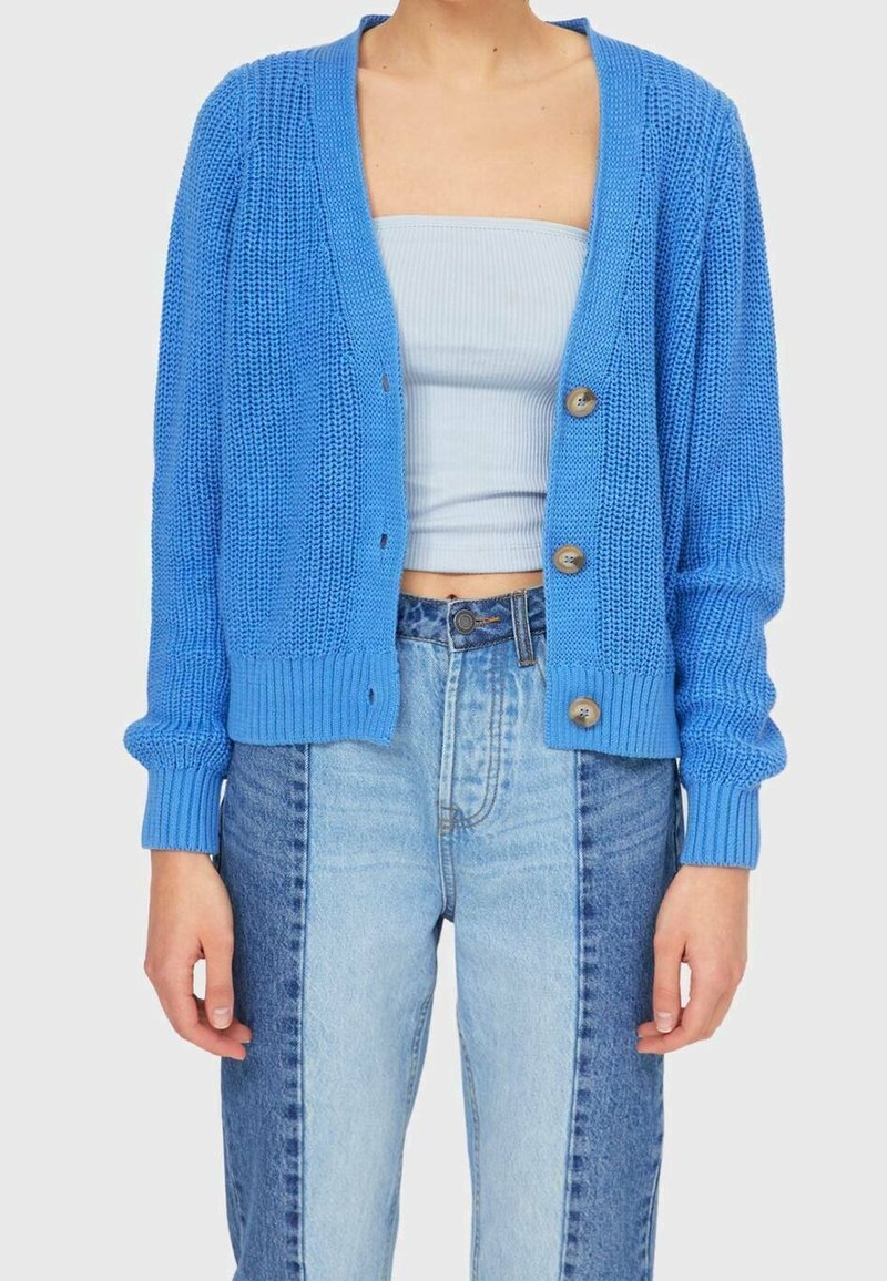 Femme portant un cardigan en maille bleu avec des boutons sur un top bandeau bleu clair et un jean taille haute bicolore, debout devant un fond uni.
