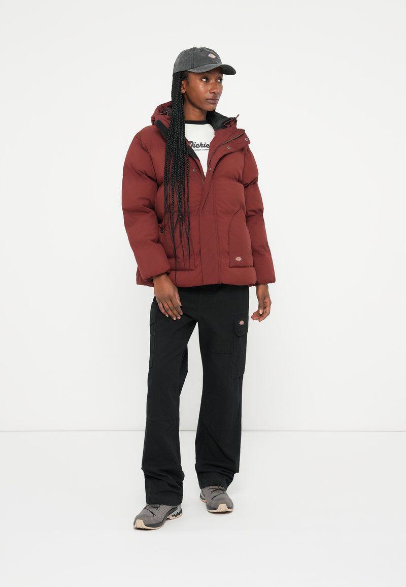 Rote Daunenjacke mit Kapuze, mit Reißverschluss und Fronttaschen. Kombiniert mit schwarzen Cargo-Hosen und einer dunklen Mütze, die den Look vervollständigt.