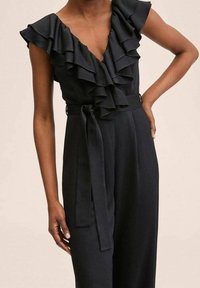 Femme portant une combinaison noire sans manches avec un col en V, des volants superposés et une ceinture nouée à la taille, posant avec une main sur la hanche.