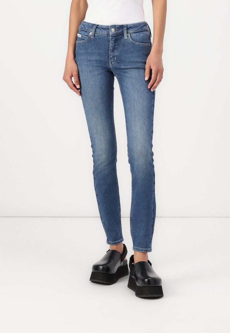 Lieknos mėlynos džinsinės kelnės su vidutinio aukščio juosmeniu, penkiomis kišenėmis ir lengvu nusidėvėjimu. Derinamos su juodais platforminiais batais.