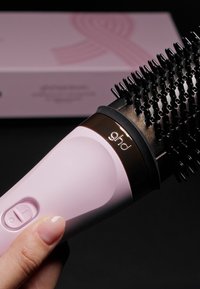 Okrągła szczotka do włosów z różowym plastikowym uchwytem i czarnym włosiem. Posiada złoty pierścień dekoracyjny oraz przycisk zasilania z boku.