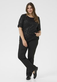 Femeie zâmbitoare purtând un top scurt cu mânecă scurtă din paiete negre, pantaloni negri și loaferi negri, stând cu o mână în buzunar pe un fundal alb.
