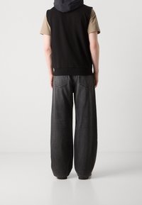 Osoba ve černé bezrukávové mikině přes béžové tričko s krátkým rukávem a širokých tmavosivých džínách, stojící na jednoduchém světlém pozadí.