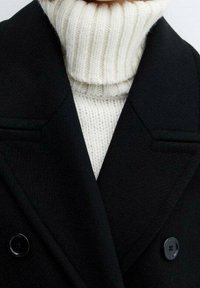 Cappotto nero doppiopetto in tessuto testurizzato, con colletto profondo e grandi bottoni, indossato sopra un dolcevita bianco a coste.