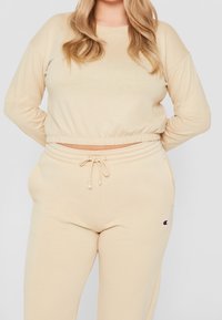 Frau trägt einen beigen, cropped Sweatshirt und passende Sweatpants mit Kordelzug und kleinem Logo am linken Oberschenkel, steht mit Händen hinter dem Rücken.