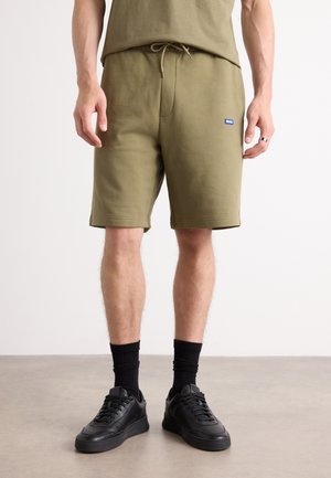 Olive green sweat shorts with an adjustable drawstring waist, side pockets, and a small blue logo patch. Black sneakers with textured soles.