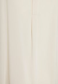Blusa beige chiaro con una texture morbida e liscia. Presenta una sottile cucitura verticale e una pattina nascosta per la chiusura. Design semplice e minimalista.