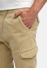 Pantalones cargo beige de una tela suave y texturizada con bolsillos laterales y traseros, que cuentan con una cintura con cordón y detalles de costura sutiles.