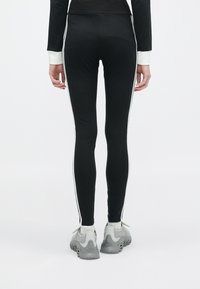 Osoba v čiernych legínach s bielymi postrannými pruhmi a šedými teniskami, stojí s uvoľnenými rukami po stranách na bielom pozadí.