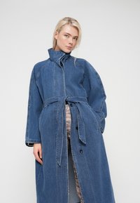Femme blonde portant un manteau long en jean bleu ceinturé par-dessus un pantalon à motifs, regardant la caméra avec confiance sur un fond uni.