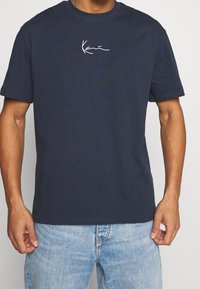 Camiseta de algodón azul marino de manga corta, con un logotipo blanco escrito a mano en el pecho. Diseño clásico de cuello redondo, ajuste relajado.