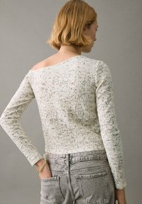 Nainen, jolla on lyhyt vaalea hius, pukeutuneena teksturoituun pois olalta tumman vaaleaan pitkähihaiseen paitaan ja kuitukankaiseen vaaleanharmaisiin farkkuihin, käsi takataskussa, kääntynyt poispäin.