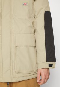 Beige jakke med svart tekstilakcent på ermet, har to frontlommer og en logo-patch. Strukturert materiale med matt finish.