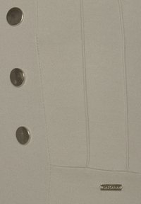 Šedá tkanina s vertikálními švy, tři lesklé kulaté knoflíky a malá obdélníková metalická visačka s názvem značky.