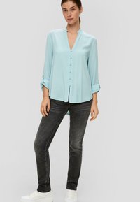 Helles Blau, langärmlige Bluse mit V-Ausschnitt und Knopfverschlüssen, kombiniert mit dunklen Jeans und weißen Sneakern. Glatter Stoff, lockere Passform.