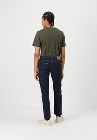 Σκούρο μπλε τζιν παντελόνι με στενή εφαρμογή, που διαθέτει καφετί δερμάτινη ετικέτα στη πίσω μέση και συνδυάζεται με πράσινο βαμβακερό T-shirt.