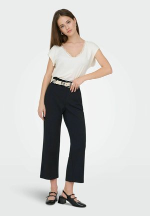 Tânără stă cu mâna pe șold, purtând un top alb cu mâneci scurte, pantaloni negri scurți, curea neagră și pantofi negri cu toc mic.