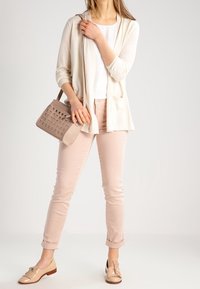 Lichtbeige cardigan met zakken over een wit shirt, gecombineerd met lichtroze skinny pants en tan instappers. Accessoire: een handtas met vierkant patroon.