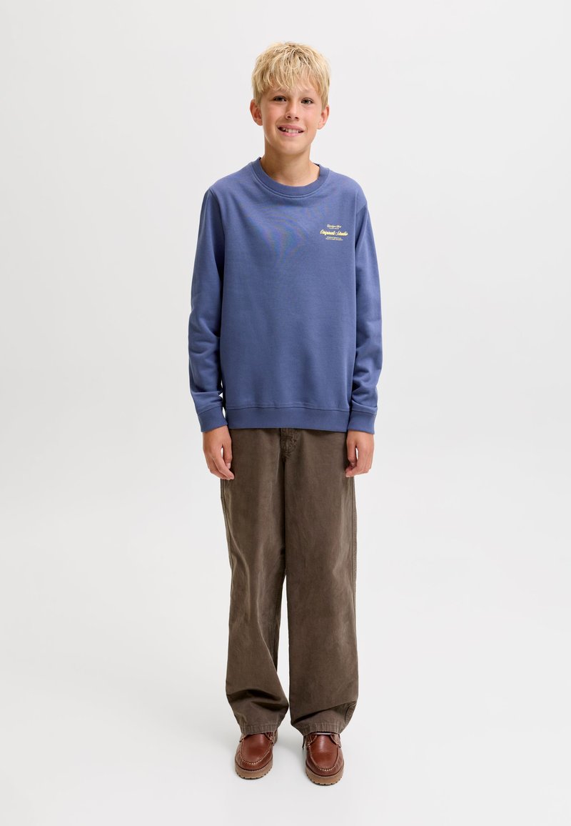 Garçon blond debout et souriant portant une chemise bleue à manches longues, un pantalon marron ample et des chaussures marron sur un fond blanc uni.