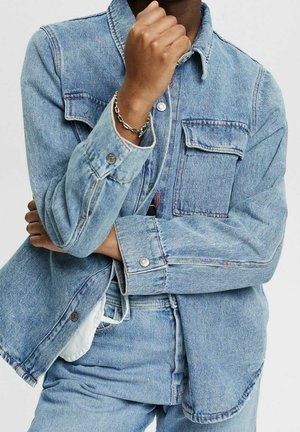 Personne portant une veste en jean bleu clair et un jean assorti, les bras croisés et la main droite près du menton, bracelet chaîne en argent au poignet.