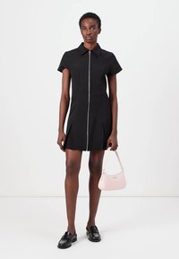 Mustat lyhythihaiset mekko, jossa on vetoketju edessä, pleattiluomus ja kaulus. Malli pitää pientä vaaleanpunaista laukkua ja käyttää mustia loafer-kenkiä.