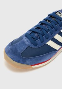 Blue sports shoe featuring a suede and nylon upper, flat navy laces, three white stripes, and a light gum rubber sole. Red and white accents.