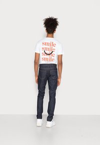 Camiseta blanca con texto rojo que dice "smile" múltiples veces; combinada con jeans de mezclilla oscura y zapatillas blancas. Se muestra la vista posterior.