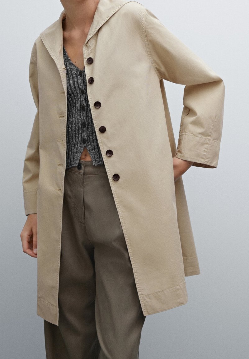 Manteau en coton léger beige avec capuche, fermeture par boutons, et manches longues, associé à un haut gris texturé et un pantalon olive ample.